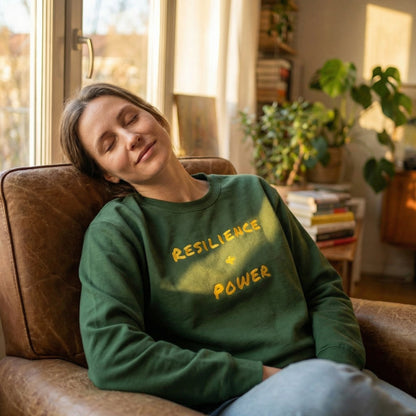 Person wearing a green sweatshirt with 'Resilience & Power' text, sitting on a couch in a cozy room.