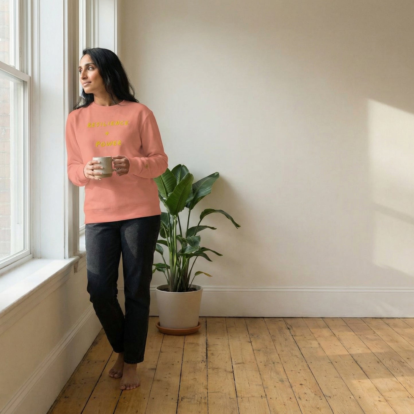 Woman in a pink sweatshirt standing by a window holding a mug, with a plant in the background.